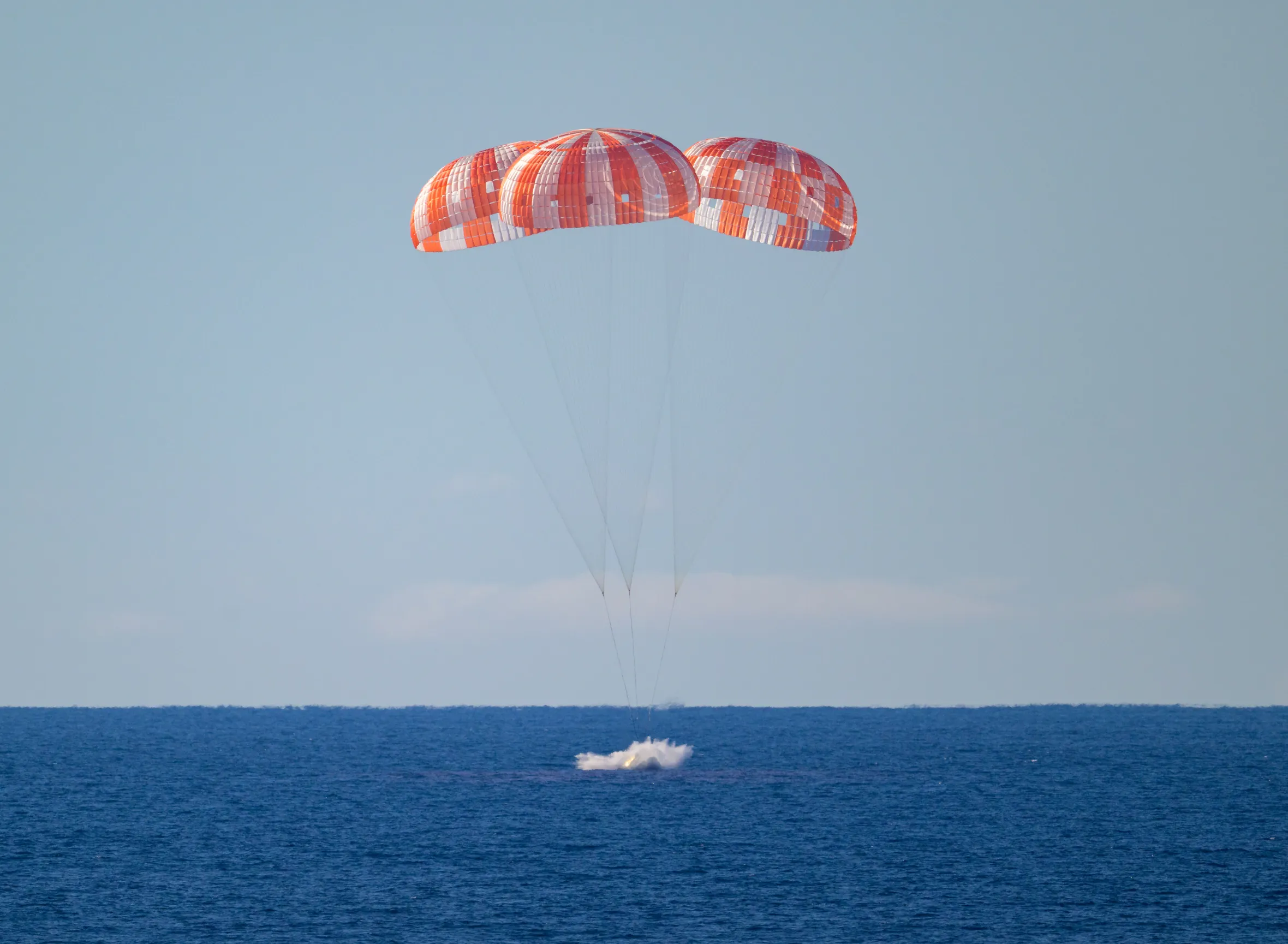 The Orion spacecraft with three parachutes above and a splash in the Pacific Ocean below.