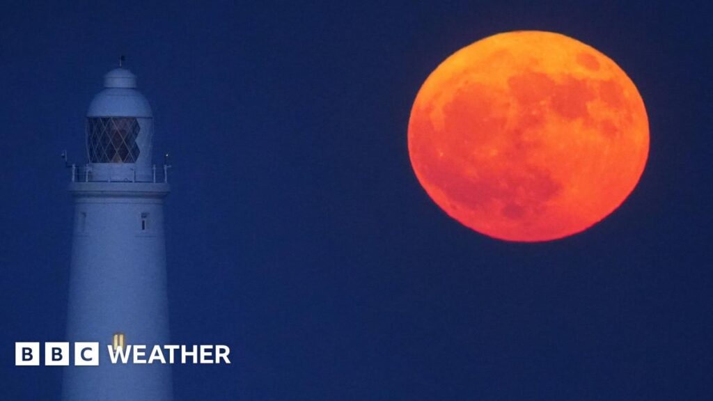 A red-orange full Moon hangs low in the night sky next to a lighthouse