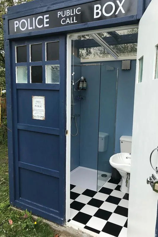 A replica of Doctor Who's time machine, converted into a bathrom. The outside is blue, beyond the open door we can see a glass shower, white toilet and a basin. The floor is chequered black and white.