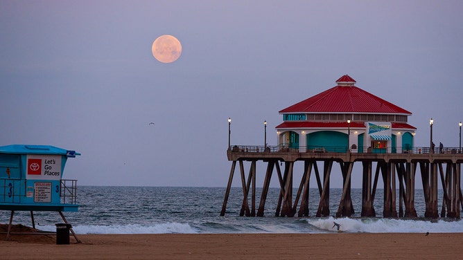 A surfer rides a wave as the full Pink Moon sets over the Huntington Beach Pier Wednesday, April 24, 2024.