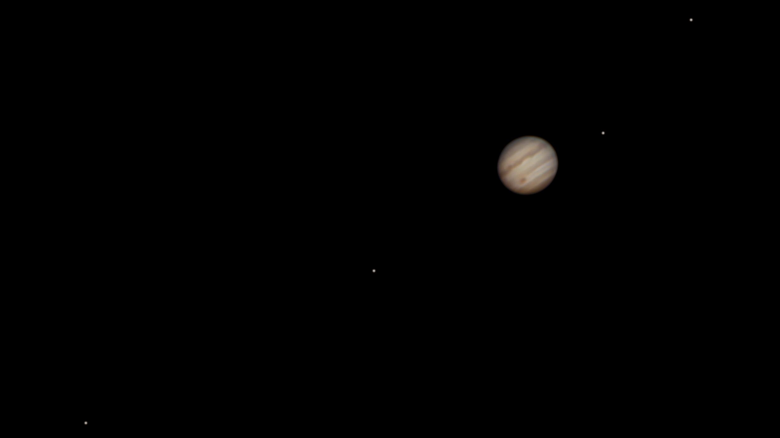 Jupiter is shown in a telescopic view in a dark sky surrounded by its brightest moons.