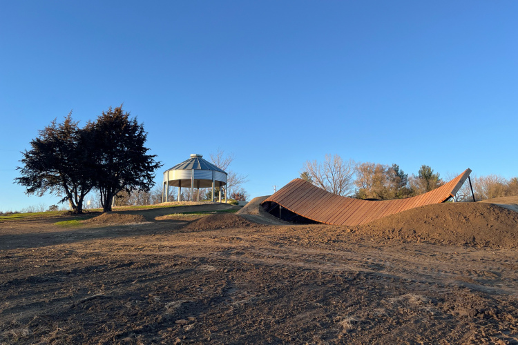 A park scene featuring a curved metal ramp and a circular gazebo on a grassy hill. The landscape is mostly bare dirt with some trees in the background, and a clear blue sky overhead.