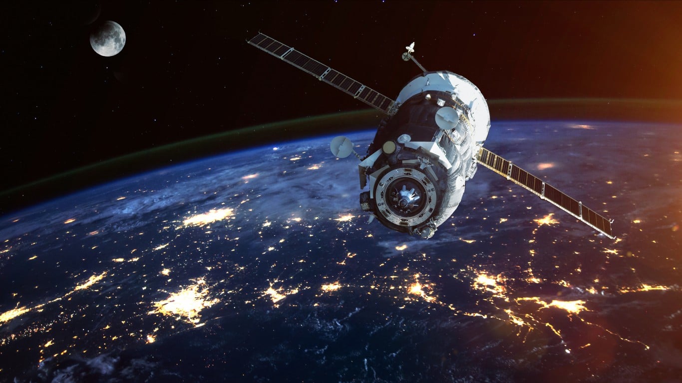 A satellite with large solar panels orbits above the Earth's night side, which is illuminated by numerous city lights. The dark expanse of space, filled with stars and a crescent moon, forms the background.