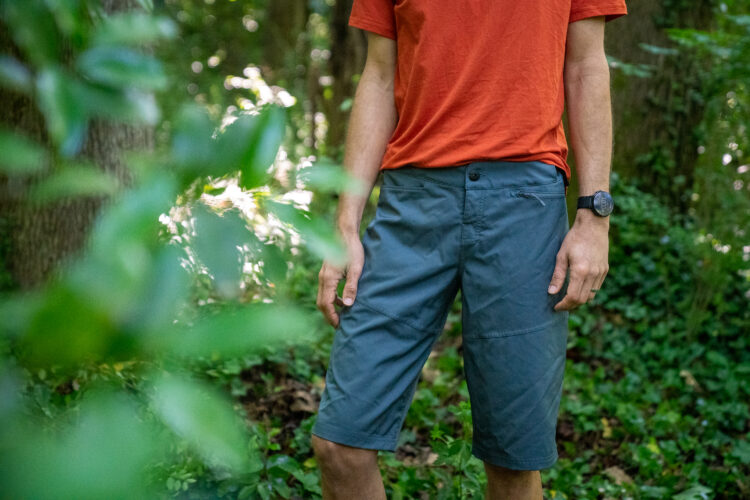 A person standing in a wooded area, wearing an orange t-shirt and gray shorts. The background features lush green foliage and trees.