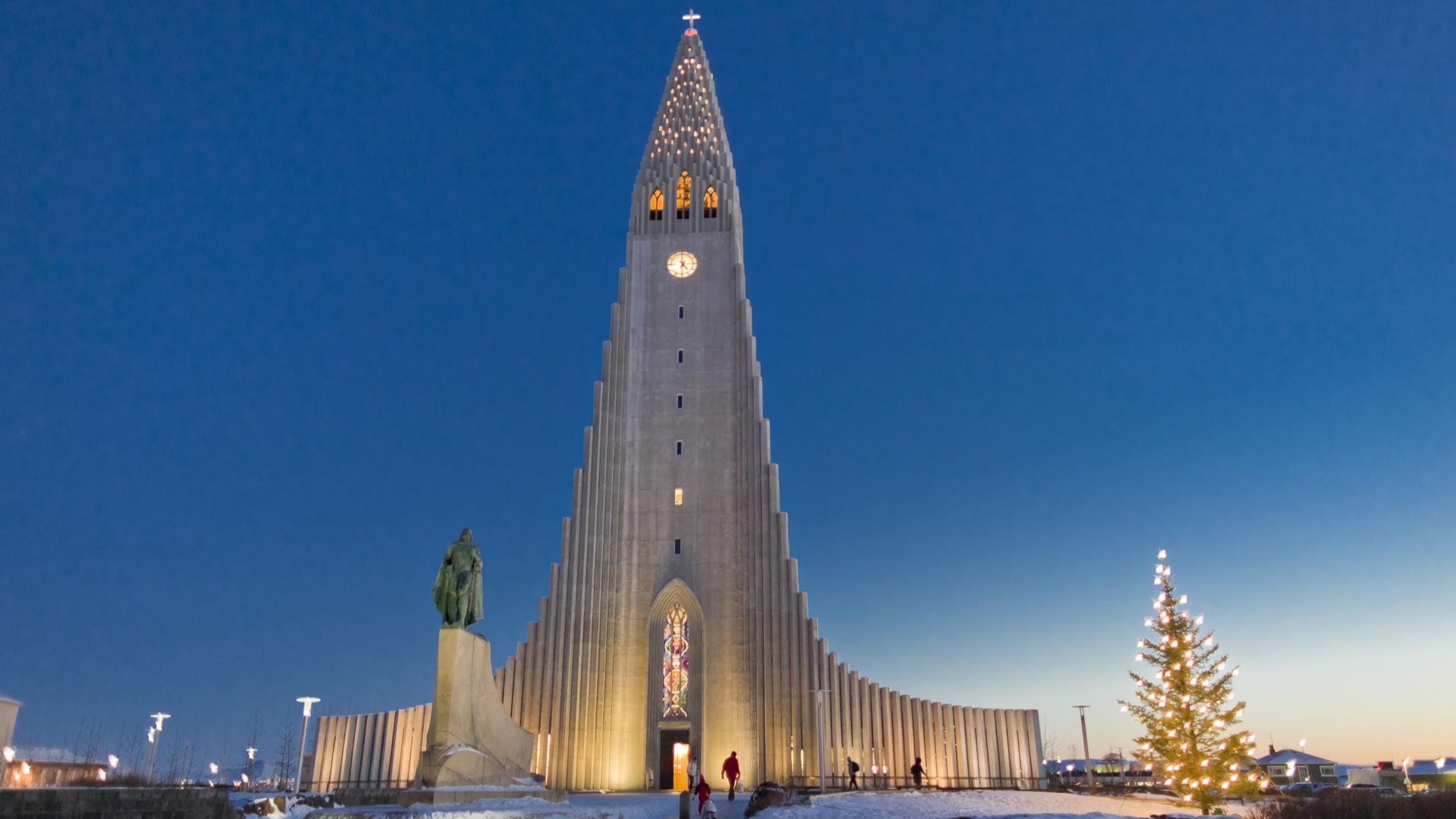 an impressive church illuminated at dusk. people walk in front of it, there is snow on the ground and a tree on the right with lights in it.