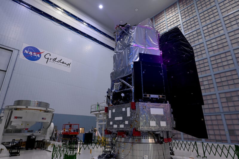 A large NASA satellite, covered in reflective material, stands upright in a spacious, high-ceilinged clean room at Goddard Space Flight Center. The NASA Goddard logo is visible on the wall in the background.