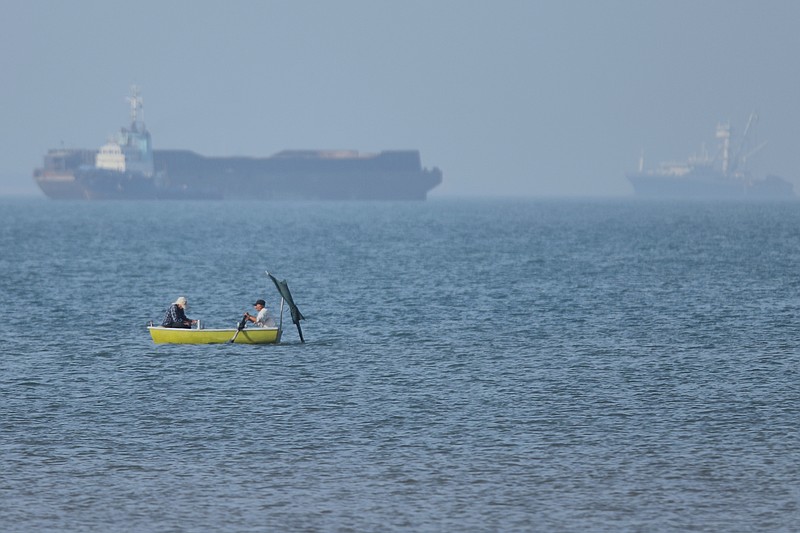 Two men sit in a small boat on the water as cargo ships are anchored in the background in the Strait of Hormuz off Bandar Abbas, Iran, Monday, May 4, 2026. (Amirhosein Khorgooi/ISNA via AP)