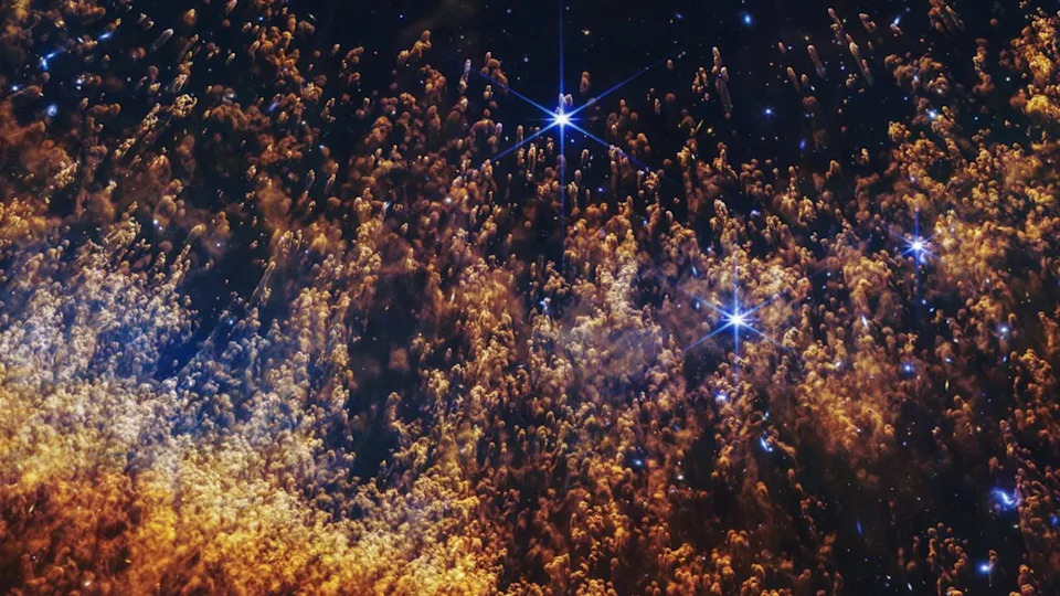 Hundreds of gold and orange clouds with feathered trails going down behind them. The small clouds are covering a few scattered, bright stars. 