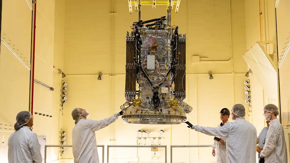 five people wearing white protective equipment stand around a rectangular spacecraft, which is hanging from the ceiling of a white-walled clean rooom