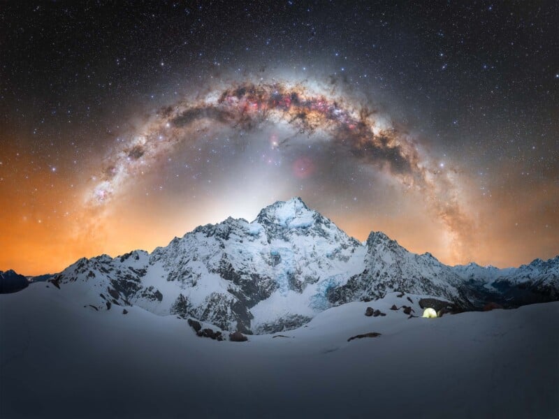 Snowy mountain peaks under a star-filled night sky with the Milky Way arching overhead. The horizon glows with orange light, and a single illuminated tent sits among the snow in the foreground.