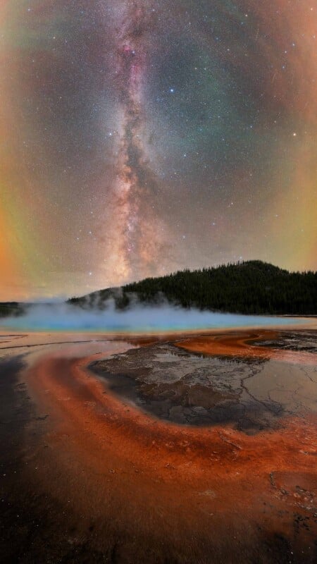 A vibrant night sky with the Milky Way arches above a steaming hot spring; the foreground features colorful mineral deposits and reflections in the water, with dark, wooded hills in the background.