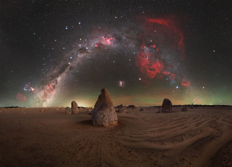 Large sandstone pillars rise from a sandy desert under a night sky filled with stars, the Milky Way arcing overhead, and bright red nebulae glowing among the constellations.