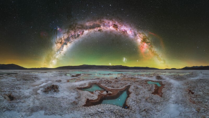 A panoramic view of a salt flat with turquoise pools under a starry night sky, featuring the Milky Way arched brightly above distant mountains.
