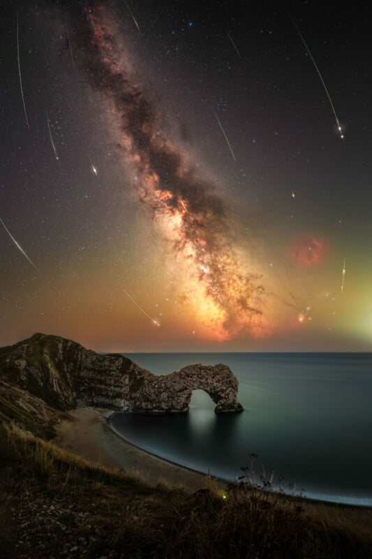 A rocky coastal arch stands by calm blue water under a night sky, with the Milky Way and multiple shooting stars visible above the horizon. Grassy cliffs frame the scene.