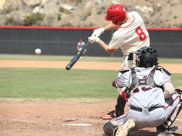 Palomar baseball player in a white uniform, number 8 swings at an incoming pitch while the opposing catcher in a grey uniform crouches behind home plate.