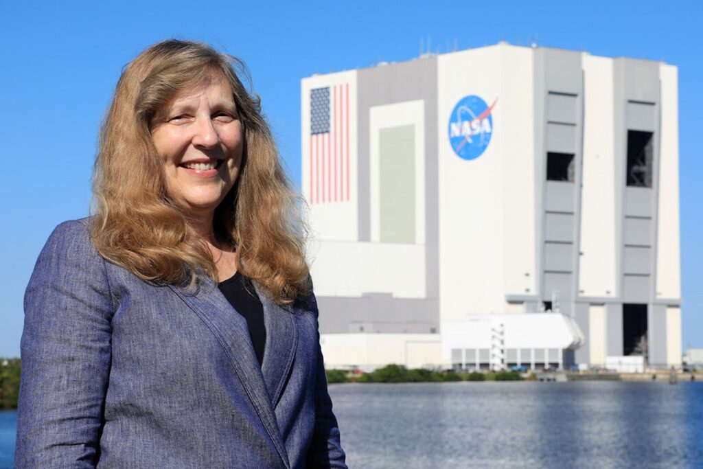 UCF Grad-Turned-NASA Engineer Focused on Sending Humans to Mars Woman with sandy brown shoulder length hair in gray blazer poses in front of square gray building with NASA logo and USA flag painted on right side