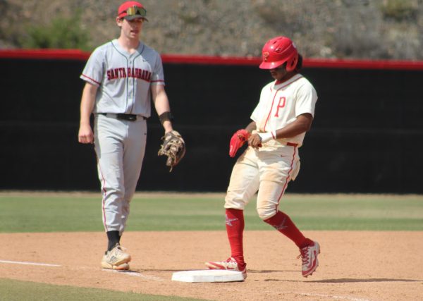 Palomar baseball player in a white uniform covered in dust steps on third base after advancing on a wild pitch, the third baseman for Santa Barbara in a grey uniform stands in the background.