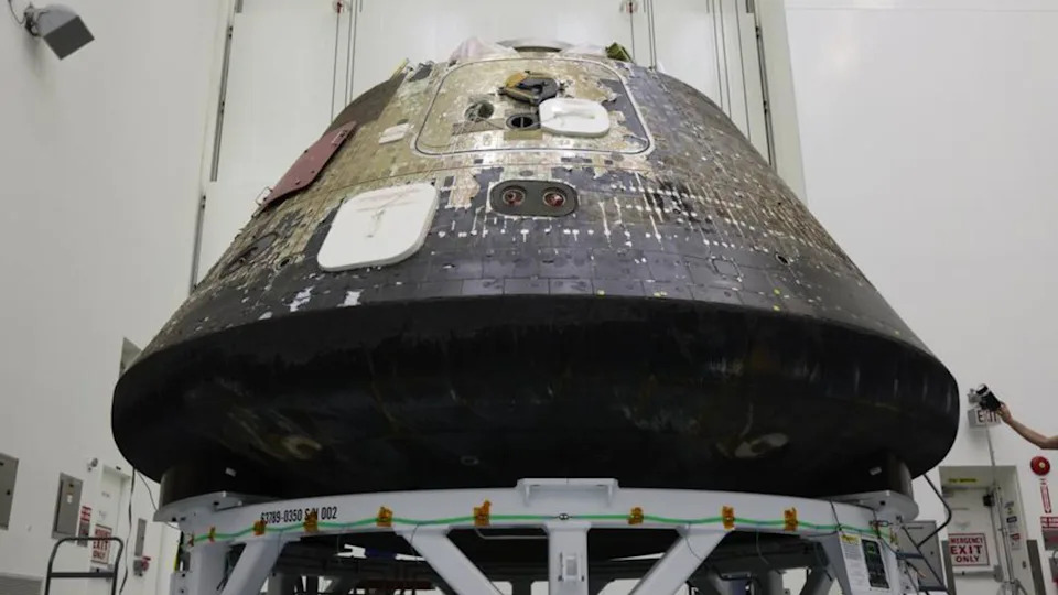 A space capsule with charred black on its bottom sits inside of a tall clean room. 