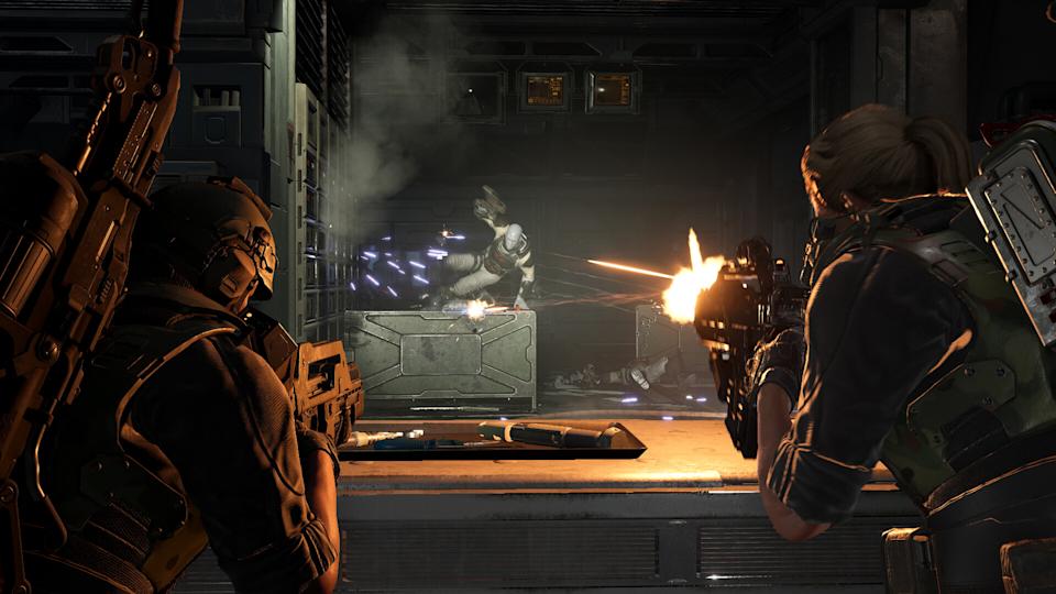 Two futuristic soldiers, one male and one female, fire at an alien creature in a dark, industrial setting. The scene is tense and action-packed.