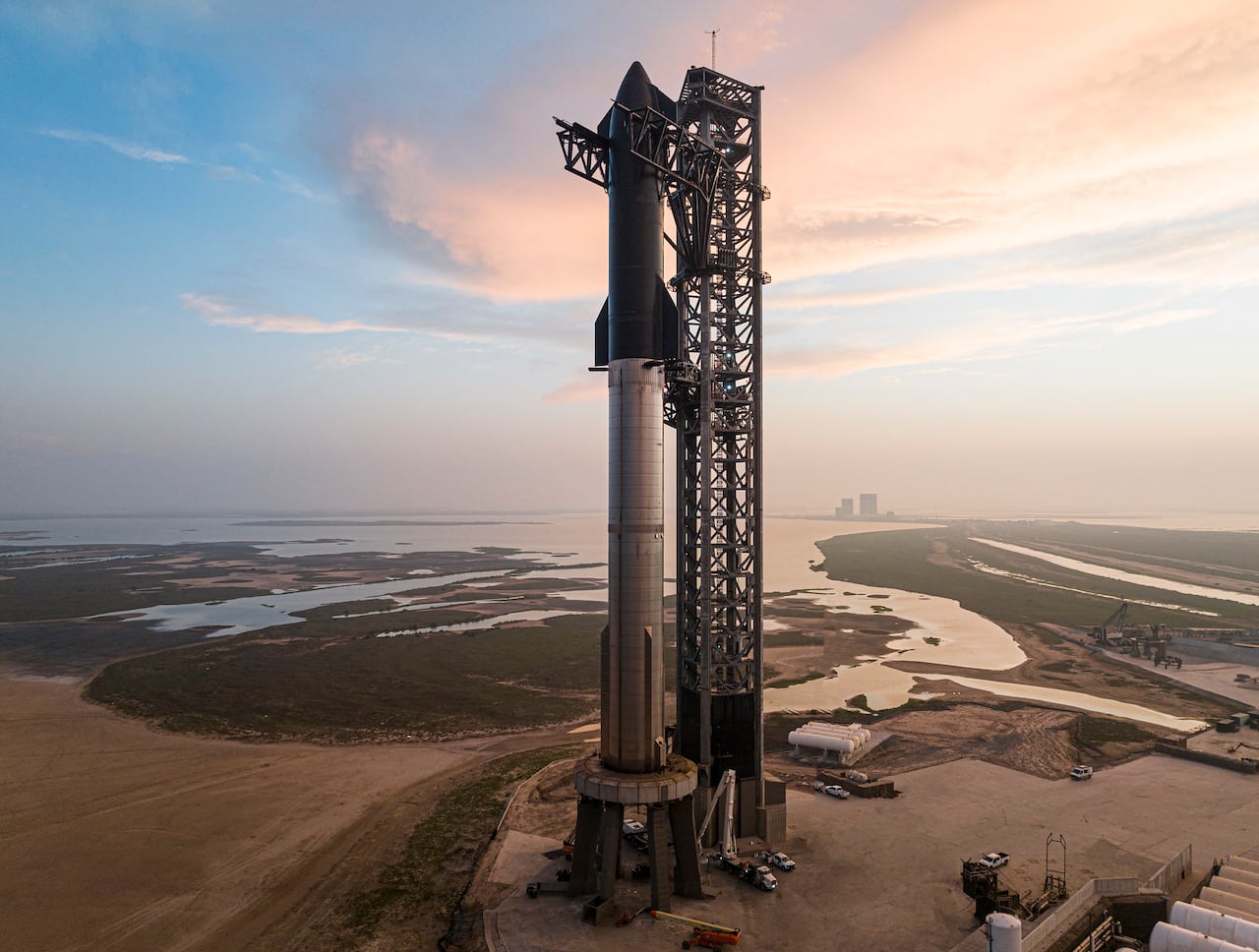 A tall rocket sits on a launch pad on a cloudy day surrounded by sand, some green grass and water.