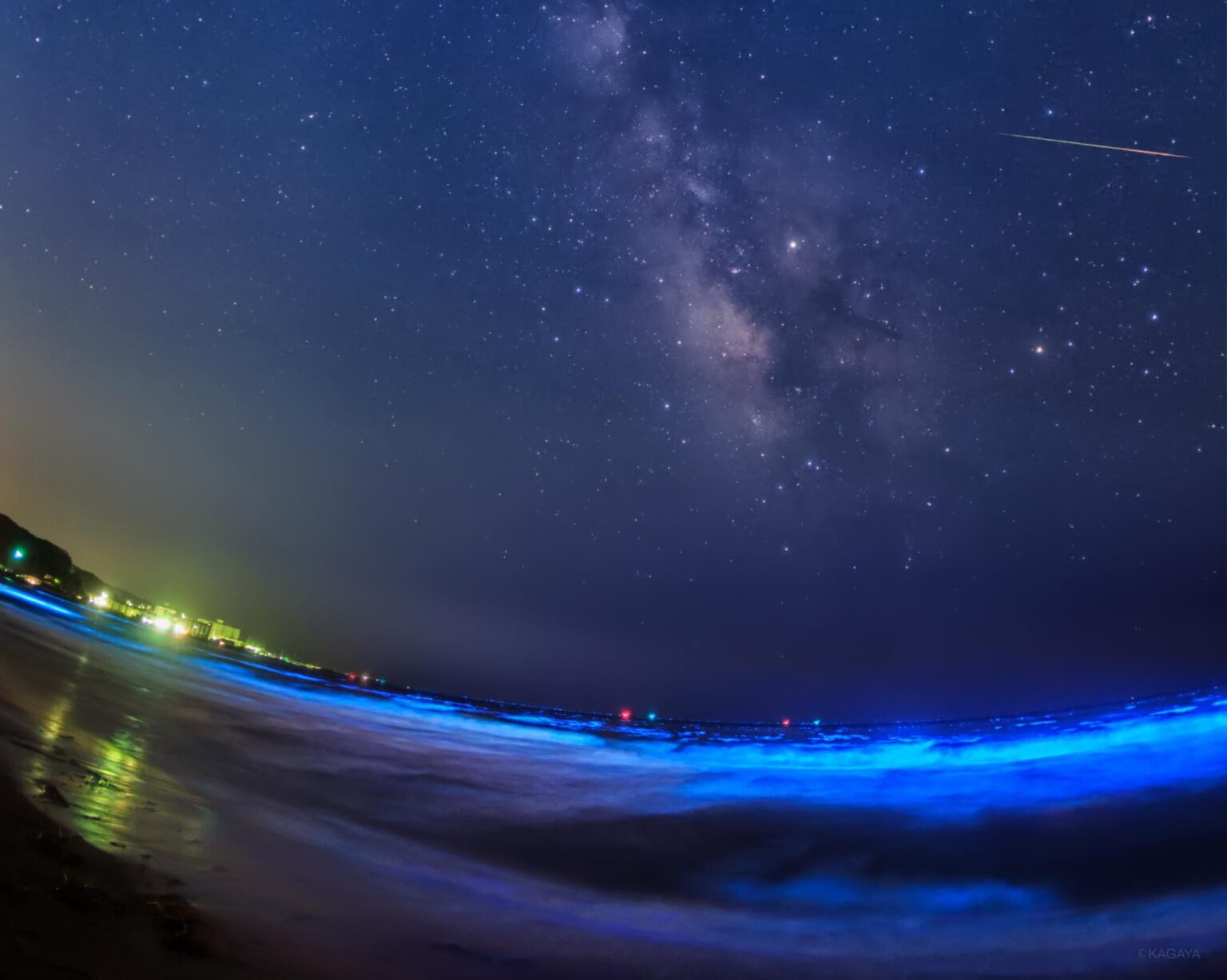 A glowing sea, the Milky Way crossed by meteor. The waves sparkle blue with the light of glowing plankton. By KAGAYA