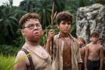 A medium close-up of two young boys standing in a tropical jungle. The boy in the foreground wears round glasses and a tan undershirt with suspenders. The boy behind him has curly hair, wears a tan button-down shirt, and holds a long, forked wooden staff. Other boys are visible in the blurred background.