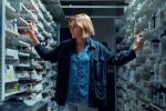 A blonde woman stands in an aisle between two rows of pharmacy shelves filled with medications. Her arms are outstretched to touch the shelves on either side.