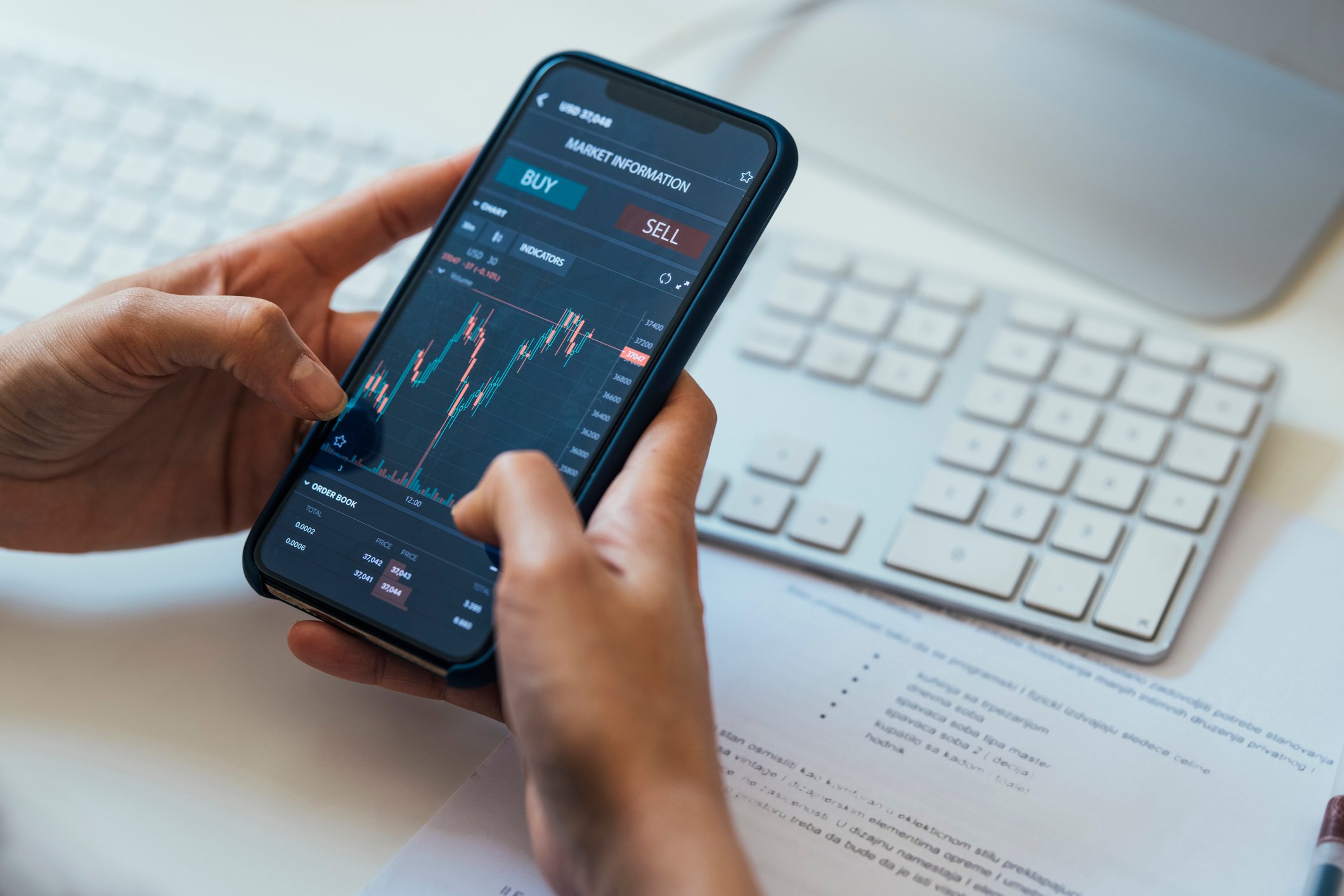 An investor holding a smartphone that's displaying a volatile stock chart with buy and sell buttons above it.