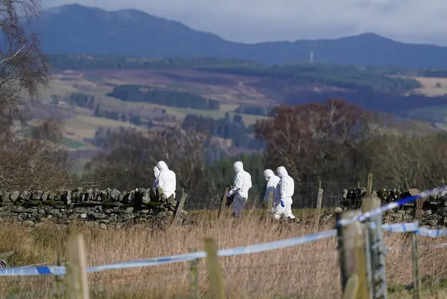 Police in white forensic suits walking in a rural area, with police tape in the foreground