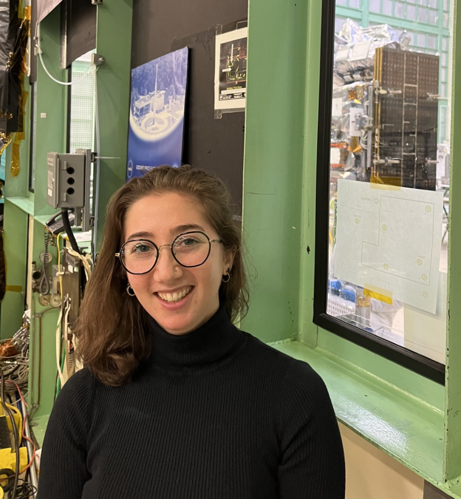 Skye Caplan stands next to a laboratory window; inside a van-sized metal instrument is visible 