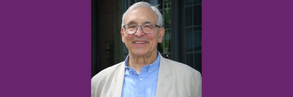 A man with white hair and glasses, wearing a cream linen jacket and blue shirt smiles at the camera