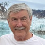 Close-up portrait of a person with short gray hair and a mustache, standing outdoors in front of a glacier and snow-covered mountains.