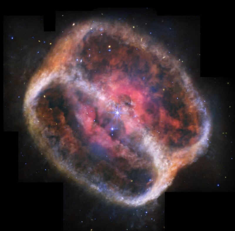 Colorful image of a nebula in space, showing a glowing, reddish-pink and purple cloud of gas with a bright central star and a large, faint, oval-shaped ring surrounding the nebula against a black background.
