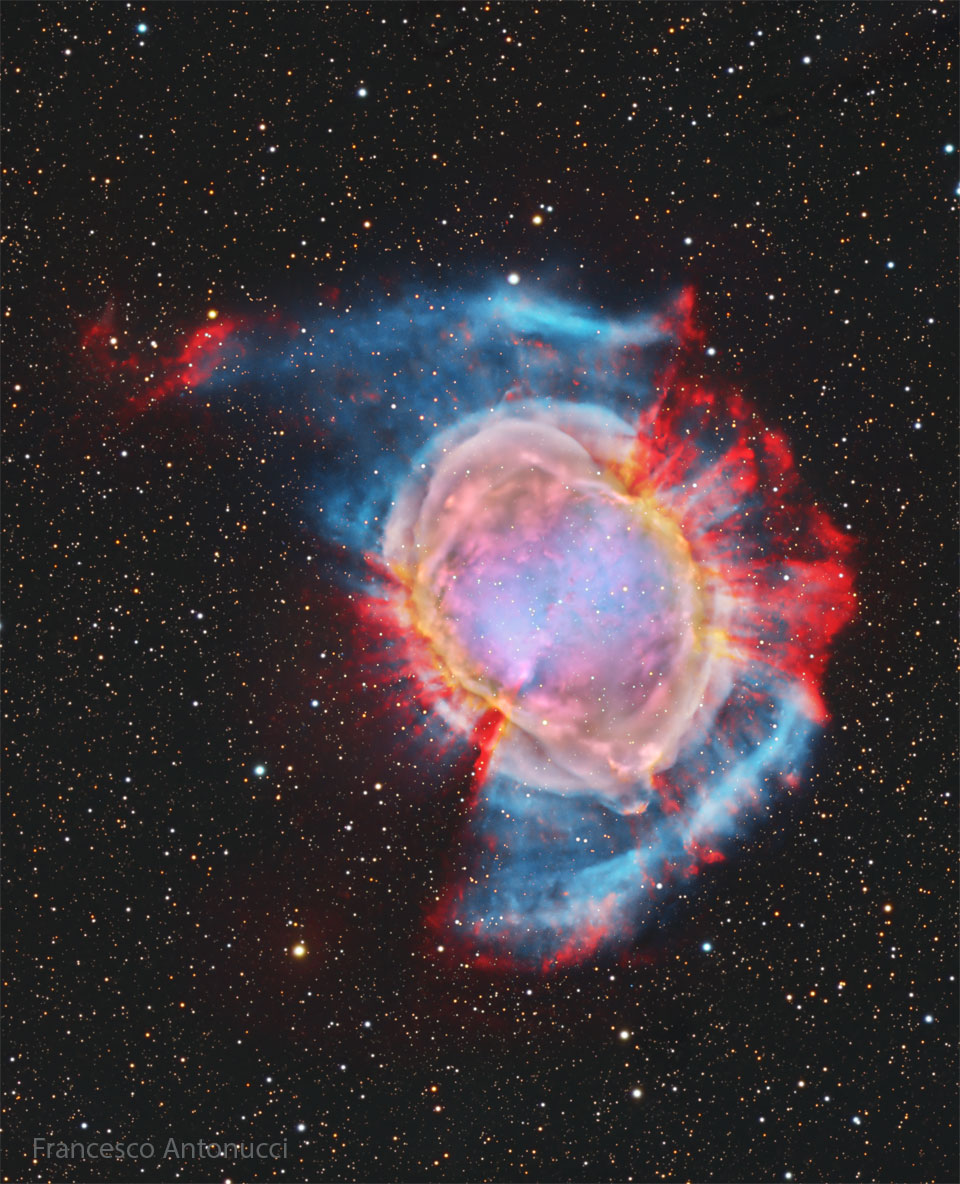 A starfield surrounds a large and colorful nebula.
Red spokes poke out from the outside of the nebula in
some directions, while a blue haze appears in other
directions. The nebula center is multicolored.
Please see the explanation for more detailed information.
