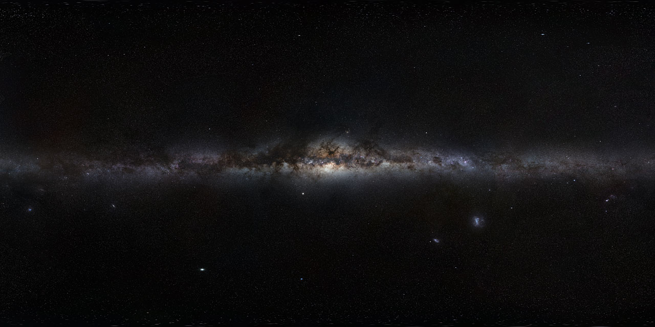 A 360-degree panoramic view of the Milky Way galaxy stretching horizontally across the frame as a bright, glowing band of stars and dust. The galactic center is visible near the middle, dense with warm orange and brown dust lanes and bright star clusters. The surrounding sky is deep black, dotted with individual stars and a few faint nebulae and satellite galaxies visible in the lower portion of the image.