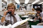 Robert Redford in a shirt and tie holds a phone to his ear as he sits behind a cluttered desk in an newsroom. Behind him, at right, Dustin Hoffman does the same, writing in a reporters notebook.