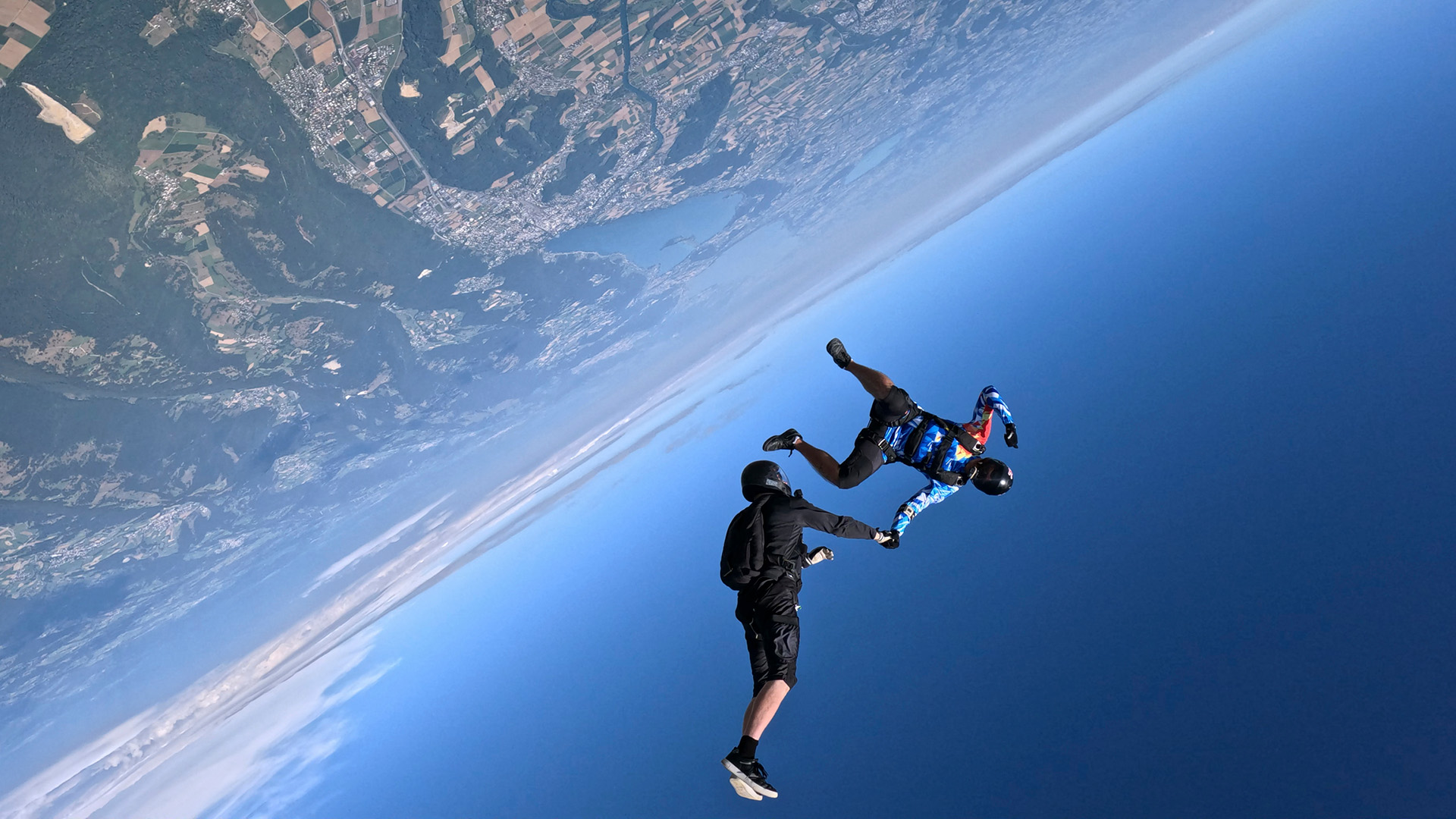 Two people hold hands while skydiving over Earth, a view of its surface next to the blue sky is seen.